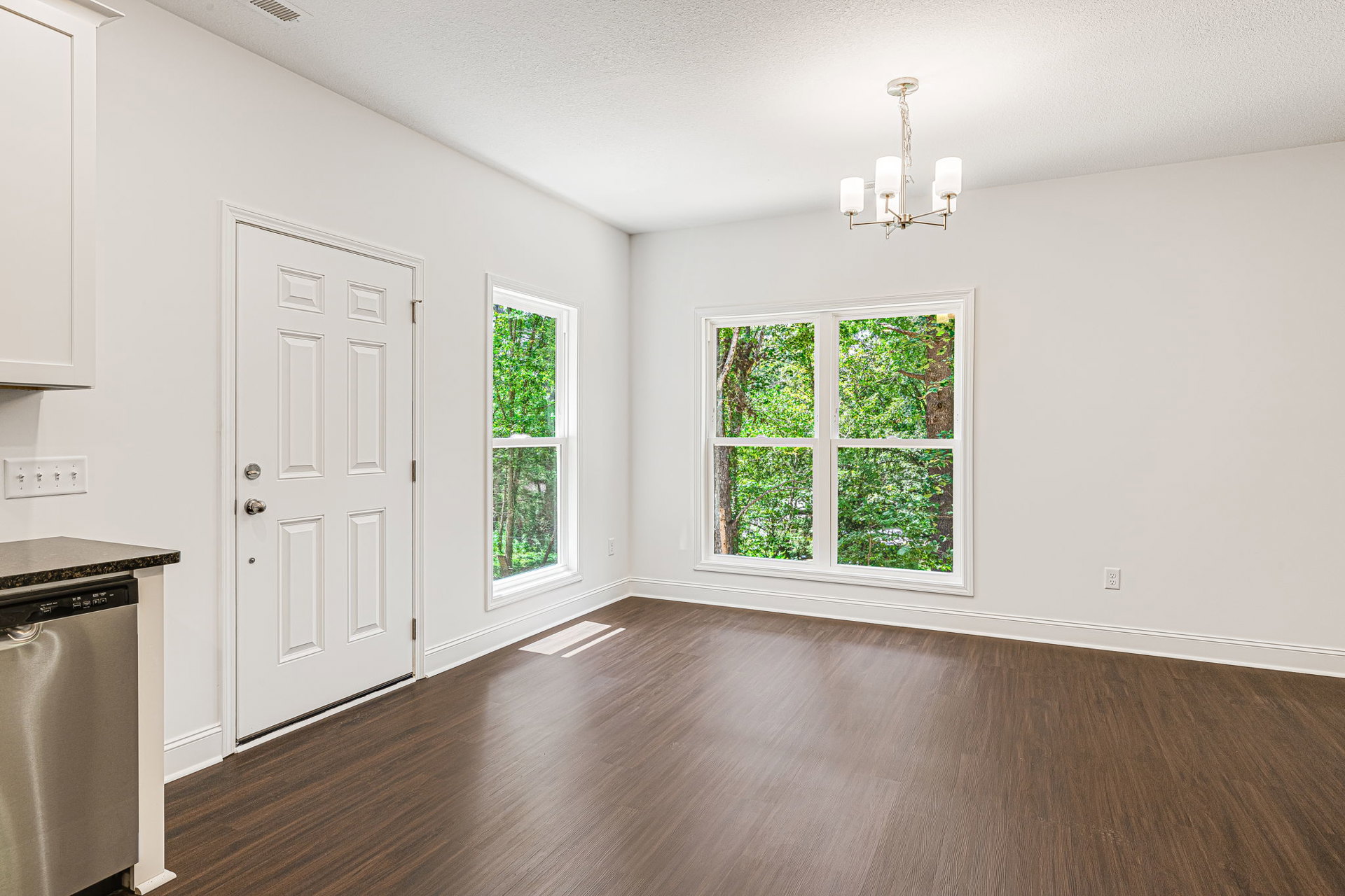 Kitchen with wood laminate flooring, white walls, silver refrigerator, white cabinetry, white door with silver doorknob, windows showing trees outside, and ceiling light fixture