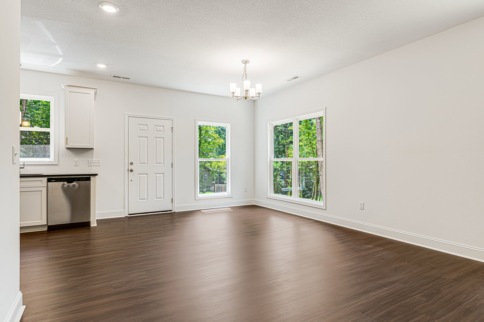 Kitchen with white walls, hardwood flooring, large windows showing trees outside, white cabinetry, stainless steel dishwasher, white door with gold knob, and chandelier overhead