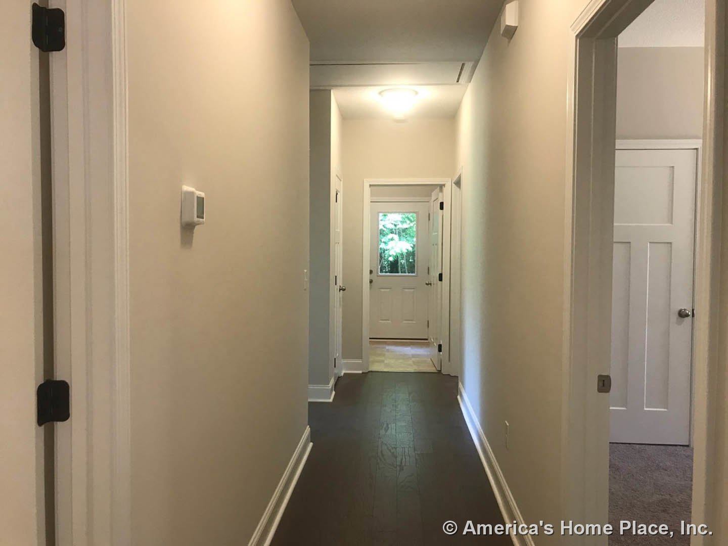 Hallway with white walls, dark wood floor, white door featuring a window and silver doorknob, white trim along the floor, rectangular wall-mounted device with a square screen