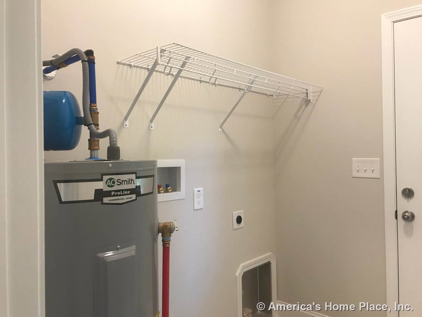 Grey tank with blue top and connected red and yellow pipes beside a white wall shelf, grey rectangular unit with logo, black circle on wall, white-framed door, and additional pipes
