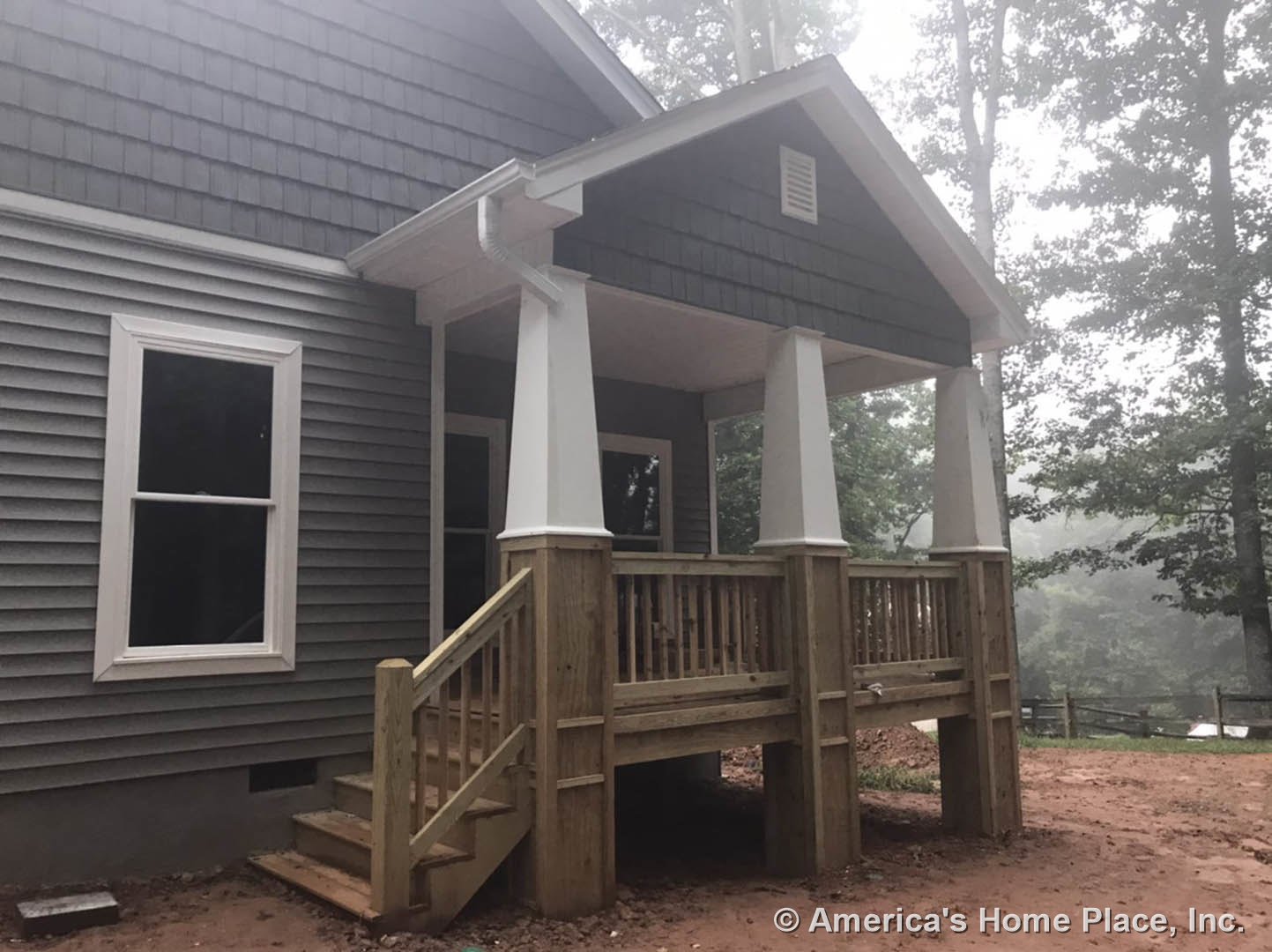 Front porch with white railings and stairs leading to wooden deck, white-framed window, vented rectangular panel on siding, roofed entry surrounded by trees and landscaped ground.