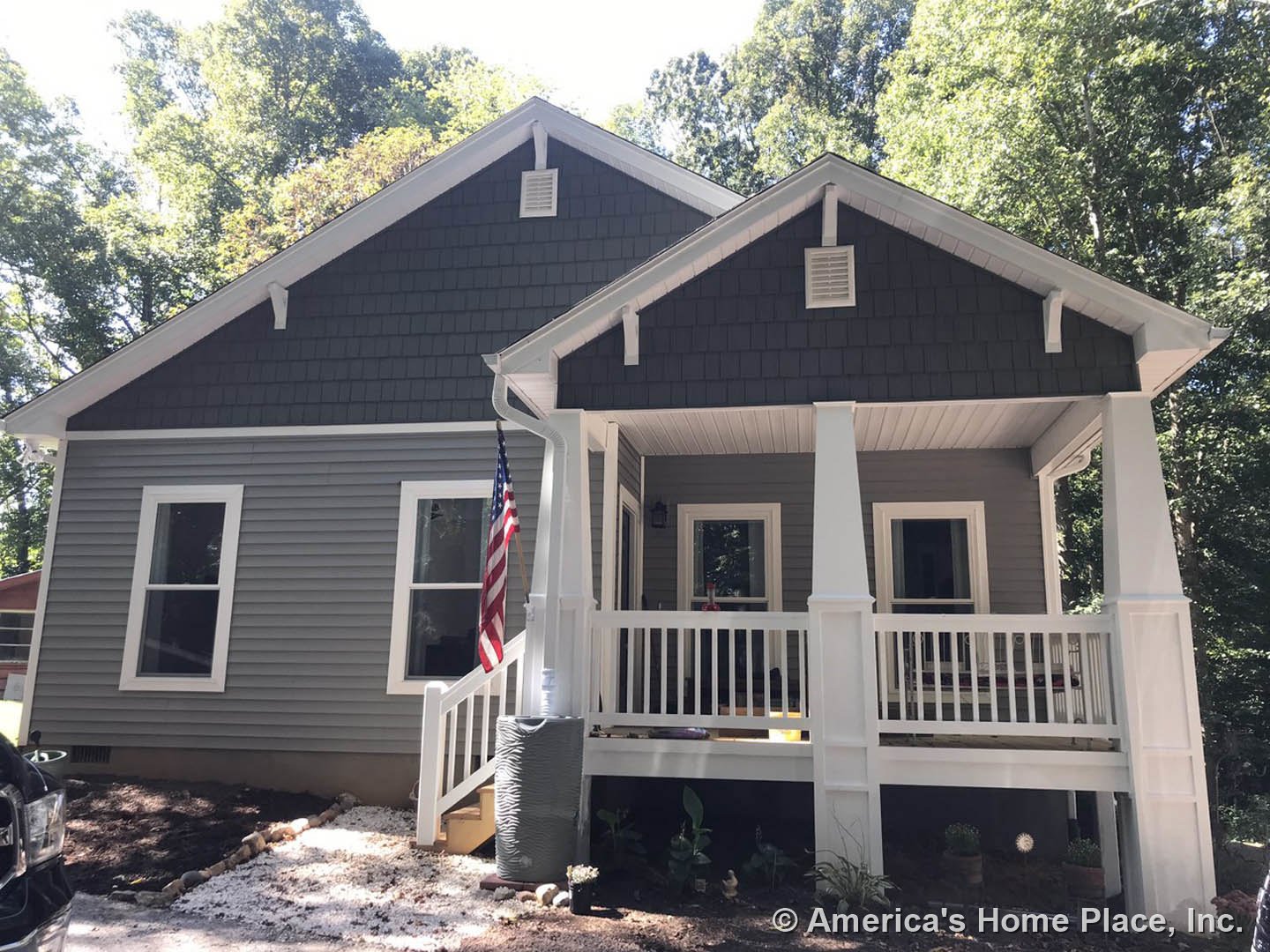 Two-story house with light-colored siding, covered front porch, American flag mounted near entrance, white-framed windows, and mature trees in landscaped yard