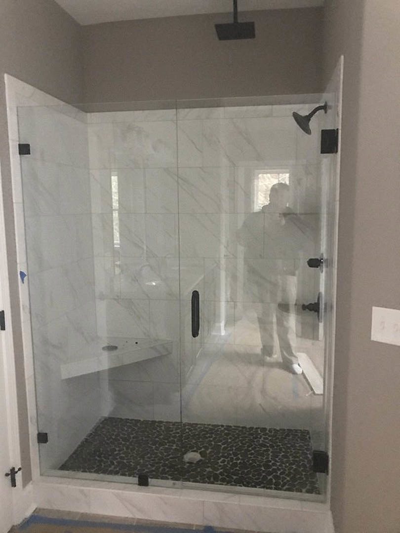 White tile bathroom with glass shower enclosure, chrome shower head, and toilet; man photographing shower near frosted window.