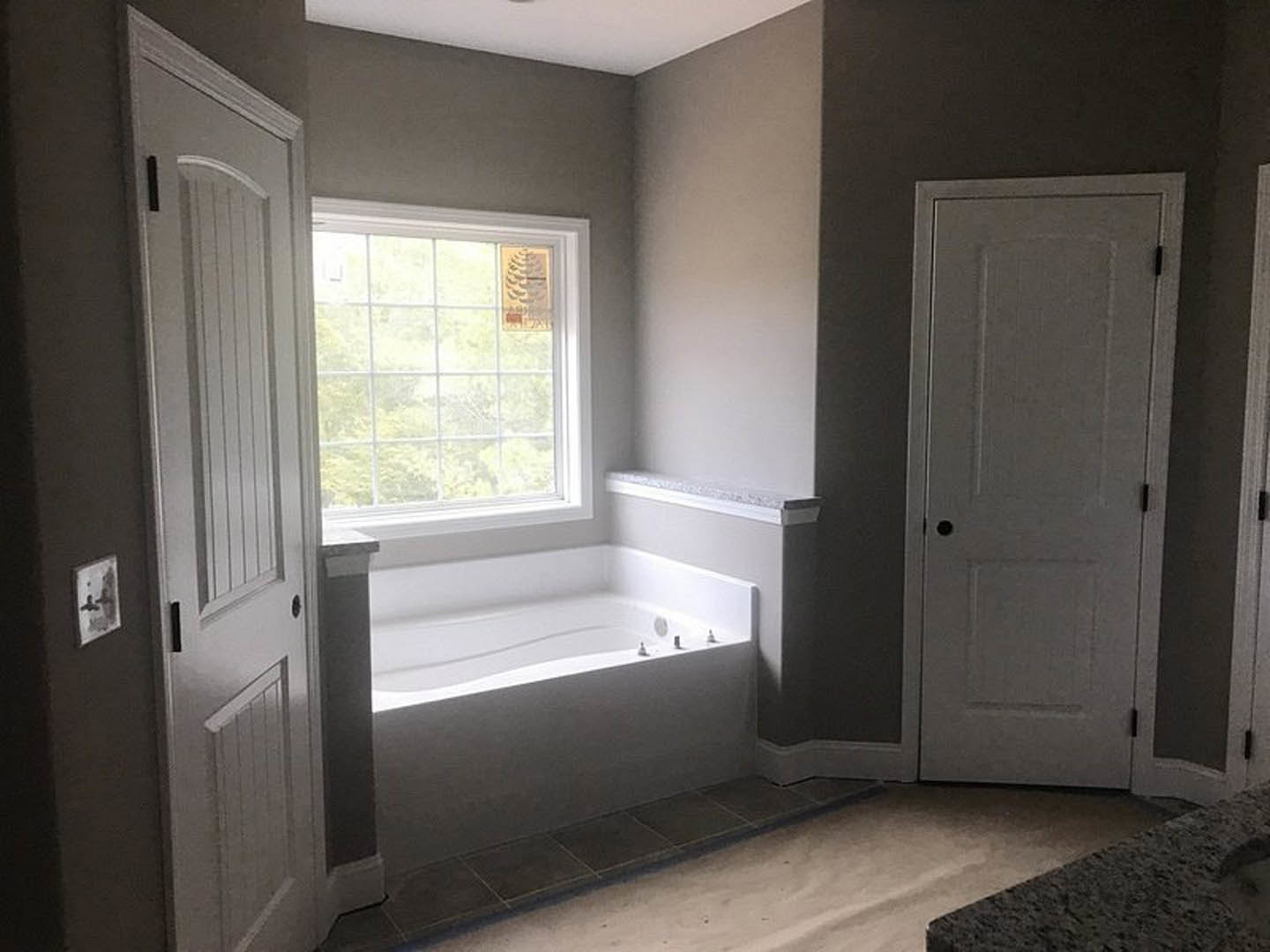 Freestanding white bathtub beneath a large window with white blinds, light gray tile floor, white walls, and black hardware on nearby door