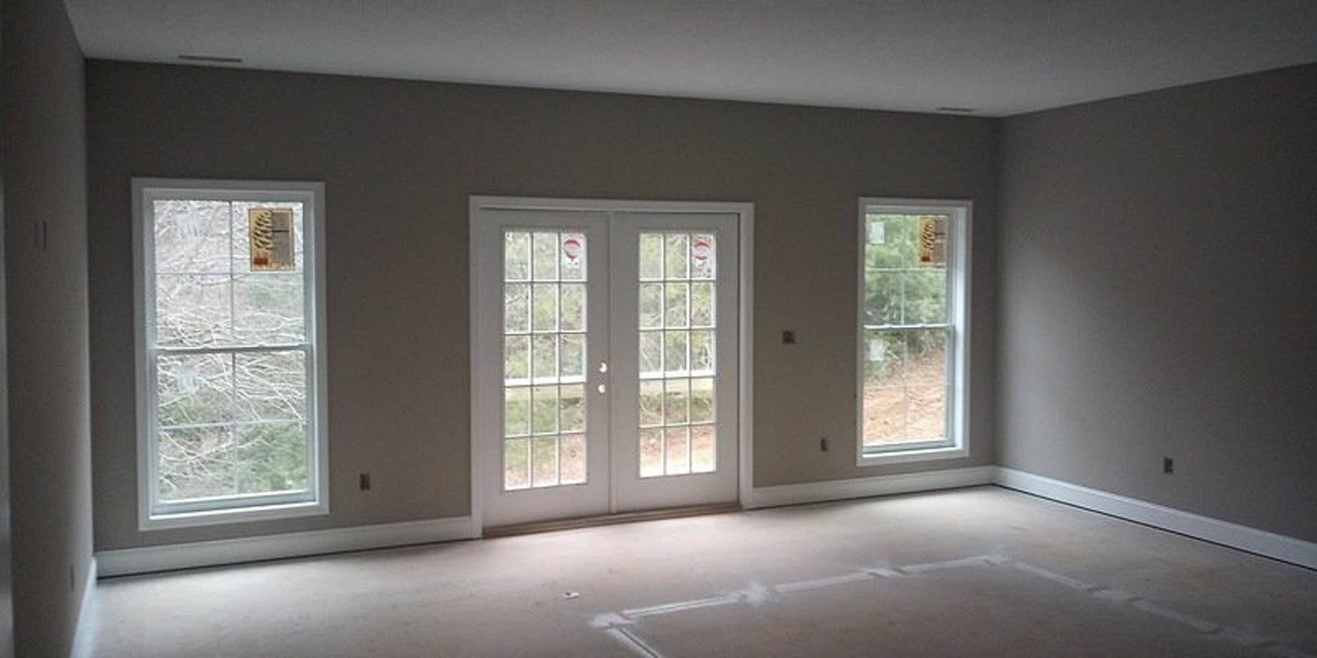 White double doors with glass panes, flanked by large windows, open onto a room with light-colored walls and hardwood flooring; outside, leafy trees are visible through the