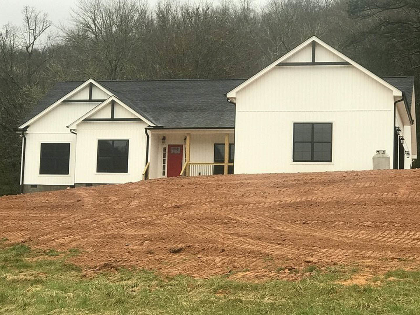 White modern house with red accent wall and square windows, set behind a dirt hill with sparse grass and small trees.