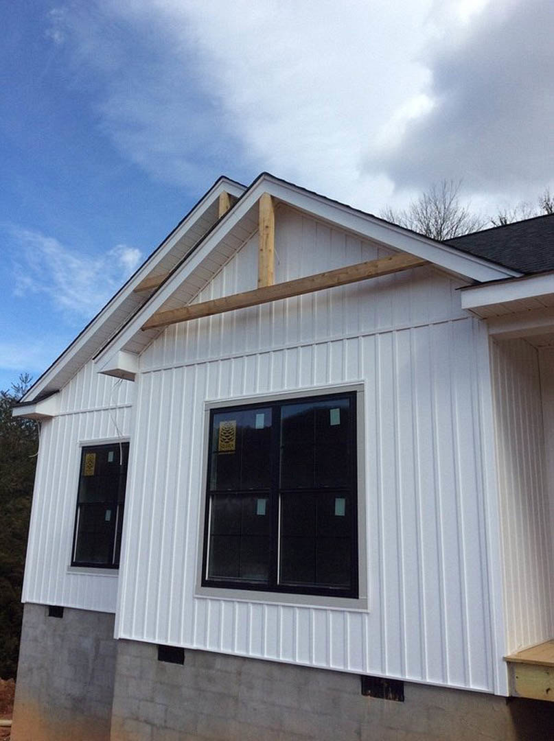 White siding exterior of a house under construction with exposed wood beams, close-up window displaying a sign, and roofline set against a blue sky with scattered clouds
