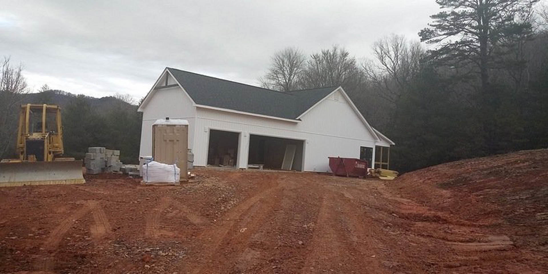 White house under construction with open garage doors, red gable roof, forklift parked near pile of white construction material, dirt road in foreground, cloudy sky and trees in