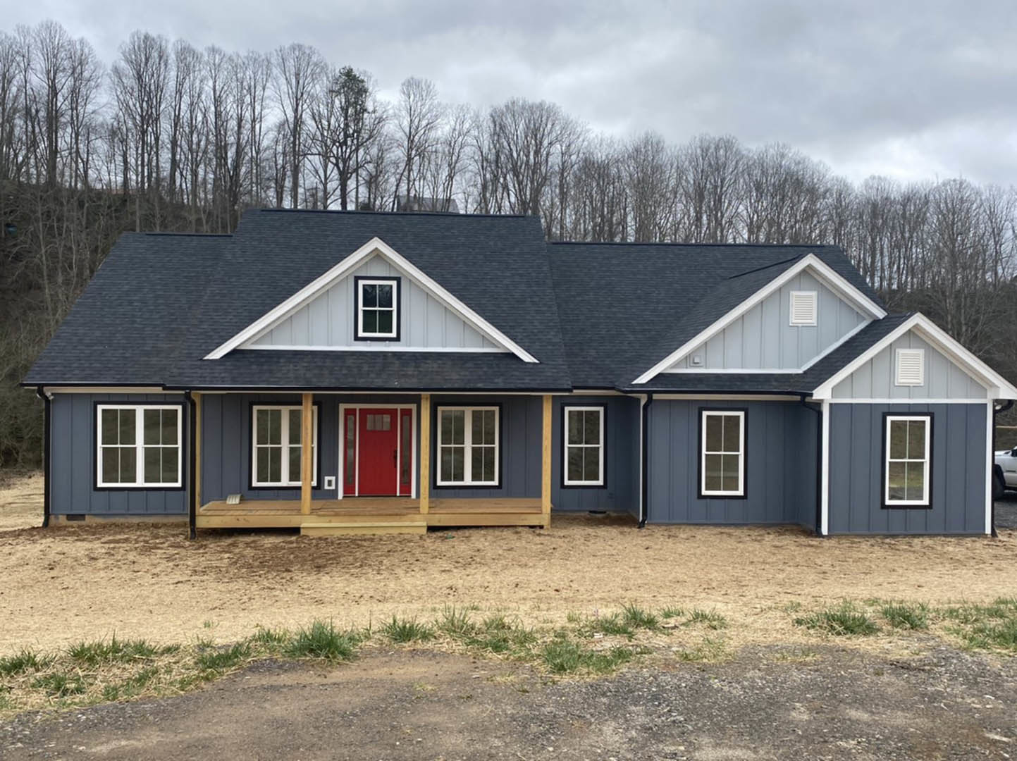 Two-story house with white siding, red front door with glass panes, white-framed windows, gray shingle roof, landscaped dirt area, and trees in the background