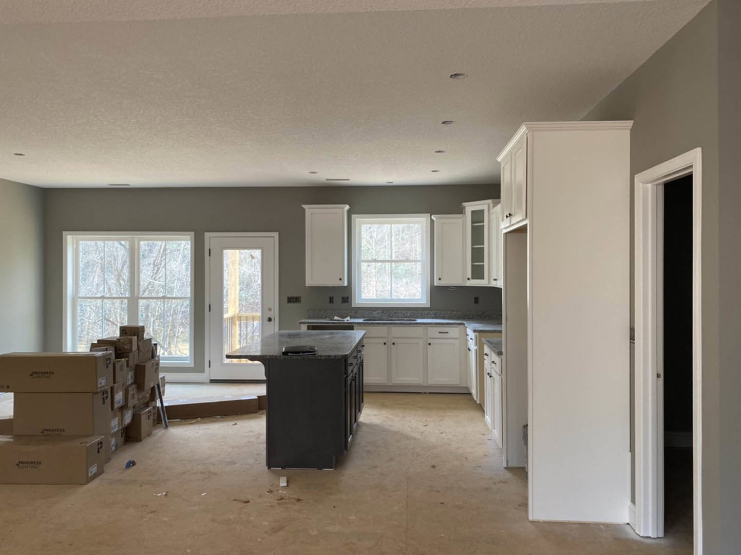 White shaker cabinets, black kitchen island with matching countertop, wide window with white trim, glass-paneled door, light wood flooring