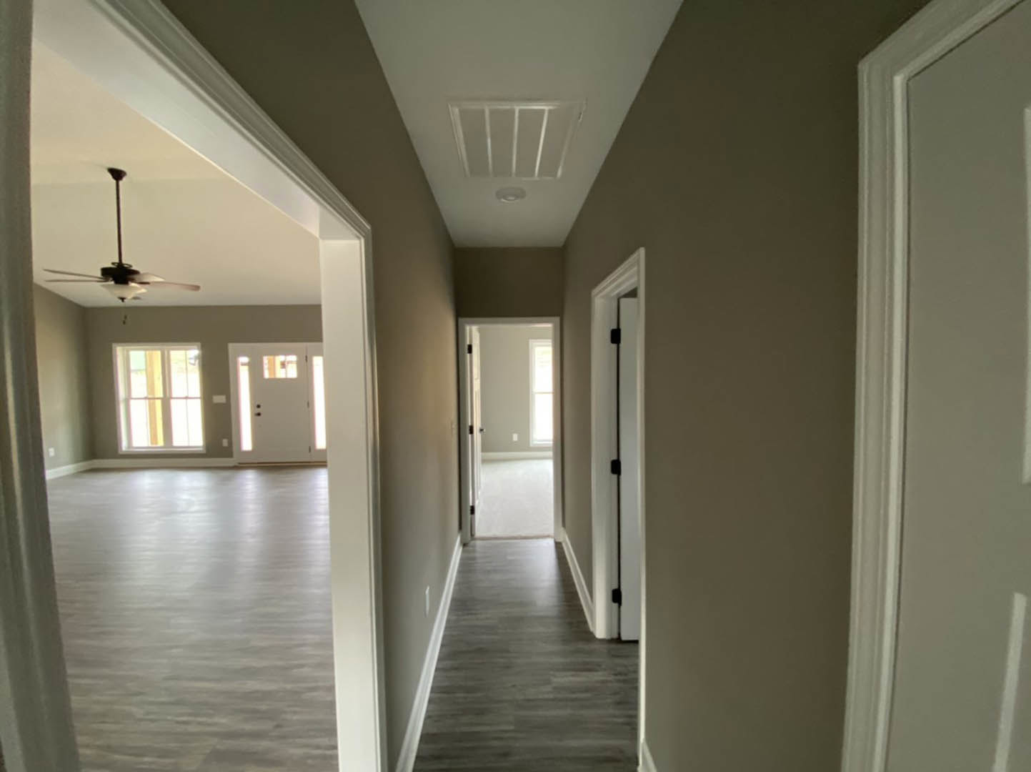 Hallway with medium-toned wood flooring, white walls, ceiling fan with light, open door revealing adjacent room, window letting in natural light, black metal door handle