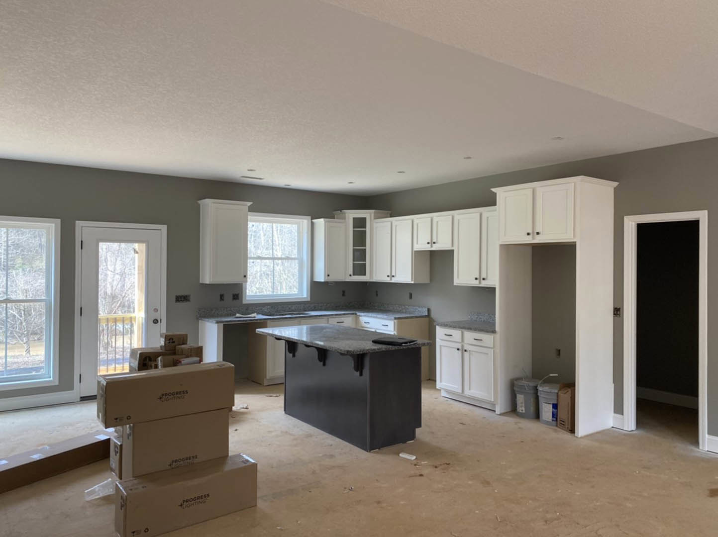 White shaker cabinets and a black island with matching countertop, stainless sink, stacked moving boxes, large window overlooking trees, glass door with white frame, light hardwood