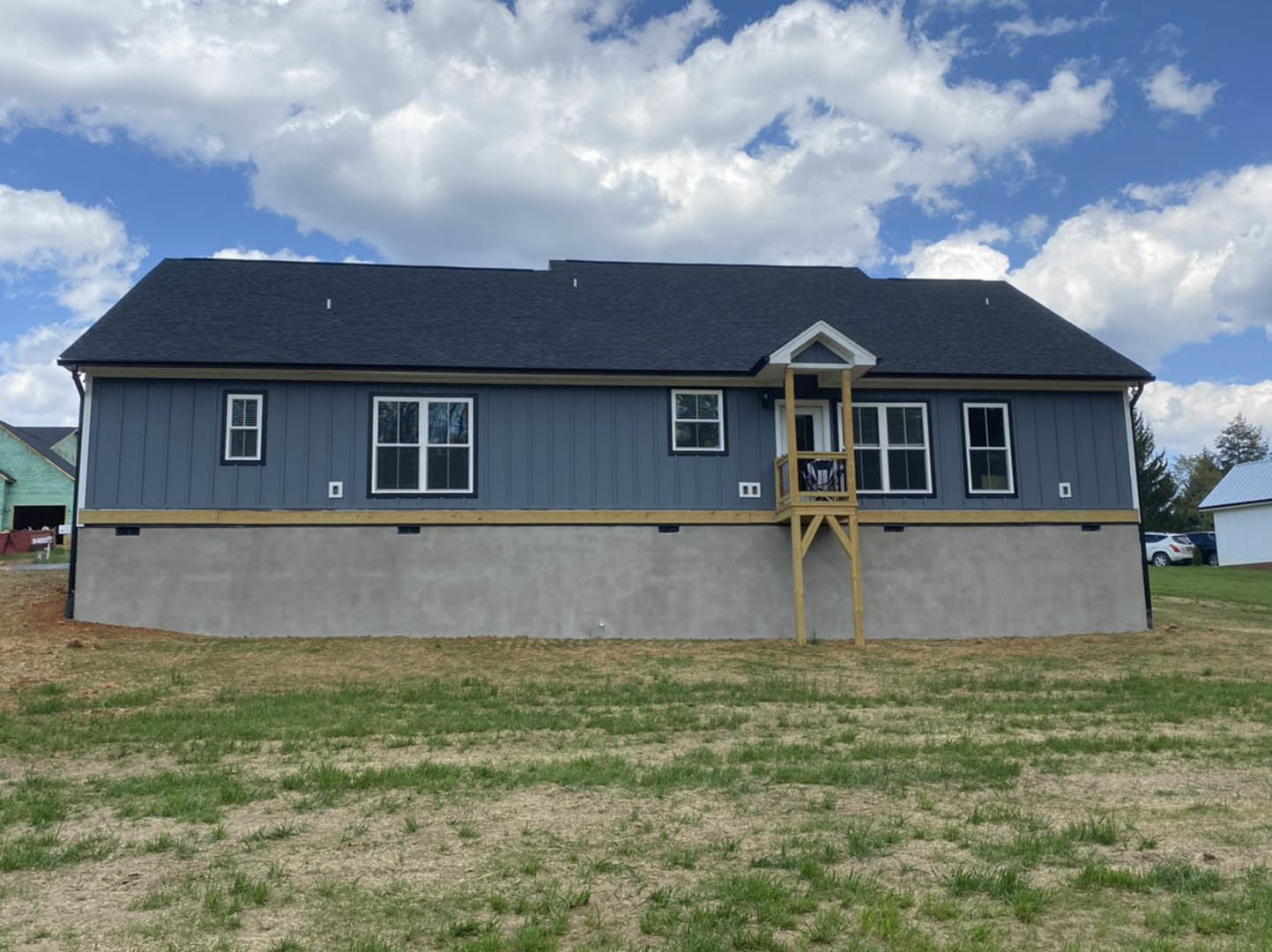 White custom home with covered porch, wooden balcony featuring a chair, large windows, surrounded by green grass field and bordered by a concrete wall under a blue sky with