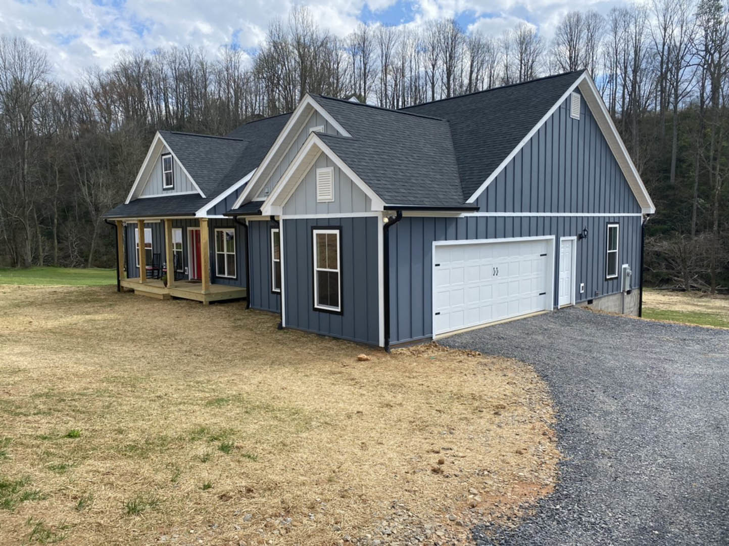 Two-story house with white siding, black window frames, covered front porch, white garage door with black handles, gravel driveway, green lawn, and mature trees in the background
