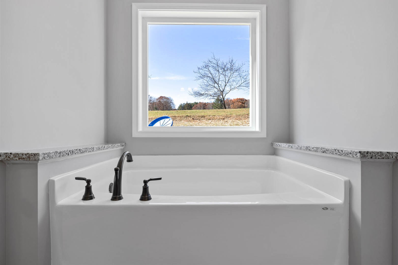 Freestanding bathtub beneath large window overlooking open field, chrome faucet, neutral tile walls, leafless tree visible outside