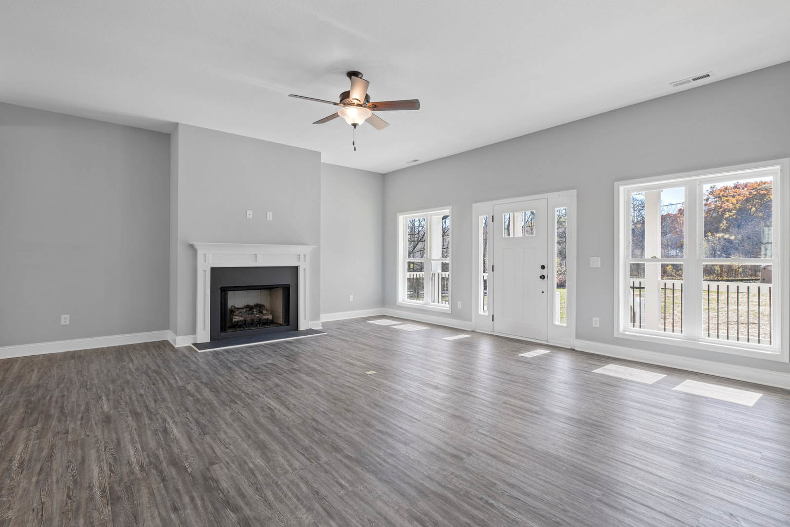 Living room with hardwood flooring, white fireplace surround, ceiling fan with light, large window showing fence and trees, and white door with glass panel