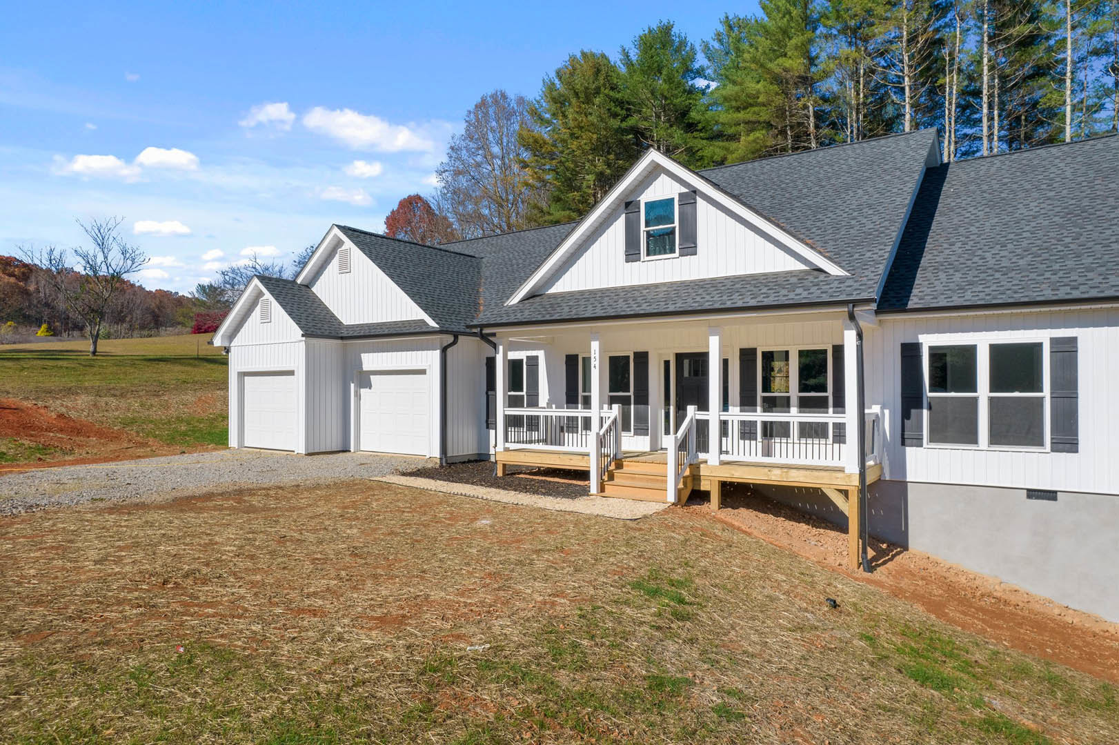 White house with covered porch, white-framed windows, concrete driveway, grassy yard, and mature trees with red leaves in the background