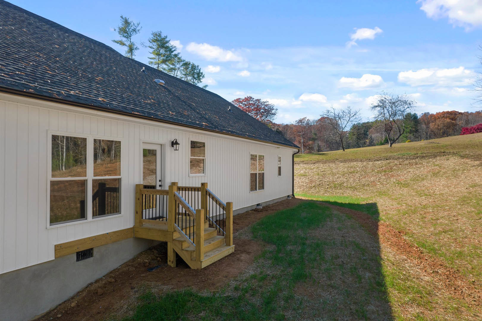 Two-story home with covered porch, wooden stairs featuring metal railings, white-framed windows, gray roof, and manicured lawn bordered by mature trees.