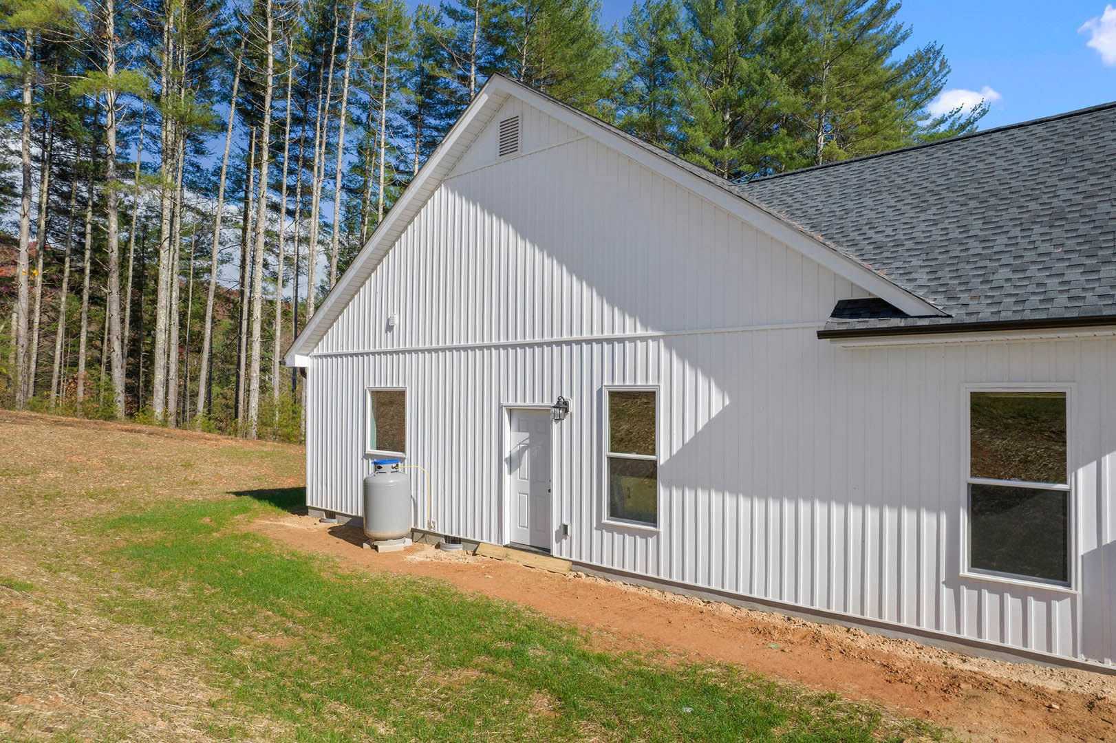 White house with horizontal siding, green manicured lawn, mature trees in background, large window with white trim, blue sky overhead