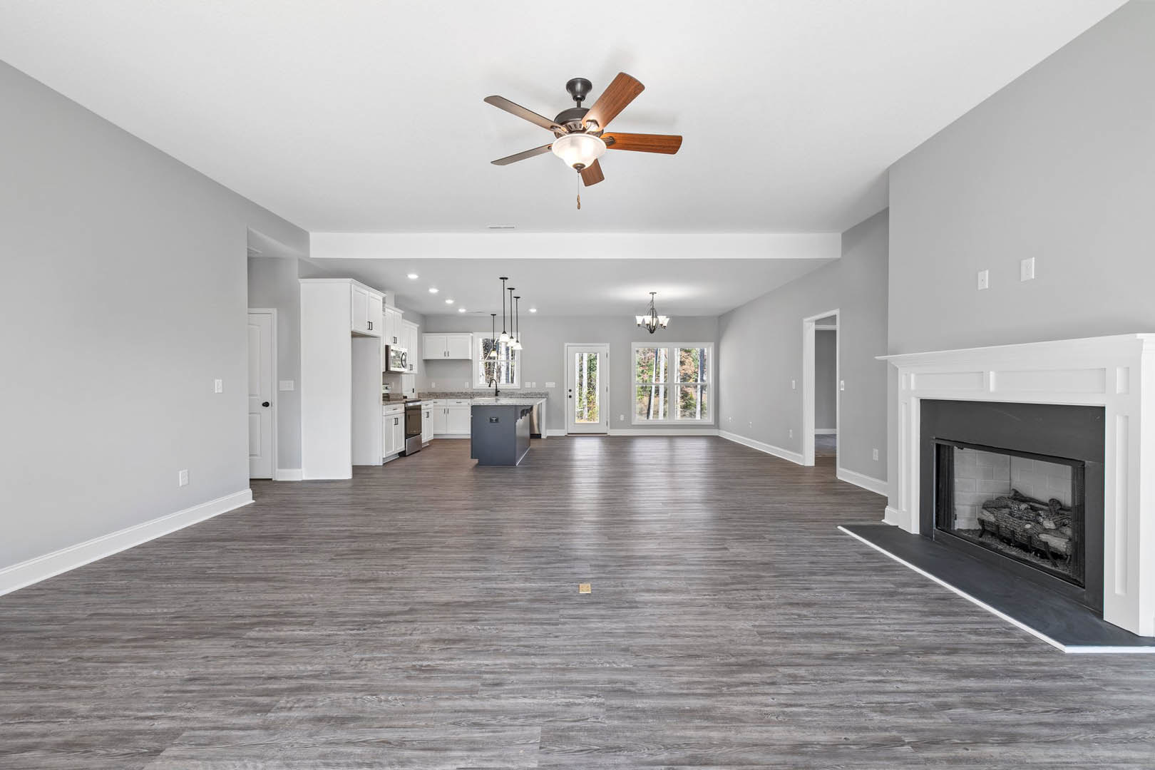 Spacious living room featuring wood flooring, central fireplace with glass doors, ceiling fan with light fixture, large window overlooking trees, and open access to kitchen area