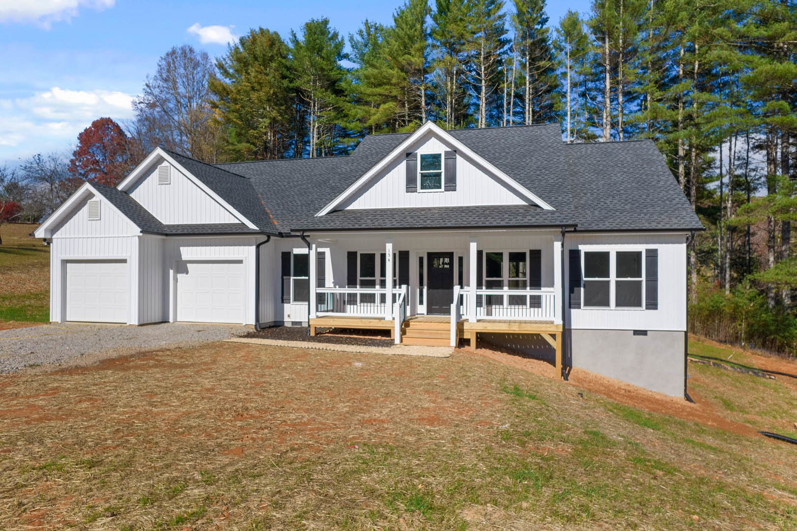 Two-story house with white siding, covered front porch, paved driveway, large windows with white frames, grassy lawn, mature trees in background