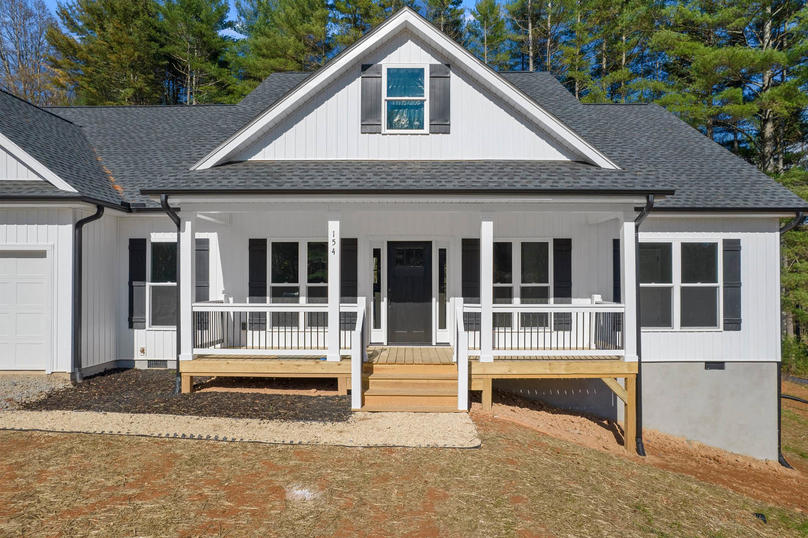 Two-story house with white siding, covered front porch, wood deck, black door with white trim, large windows, and gabled roof.