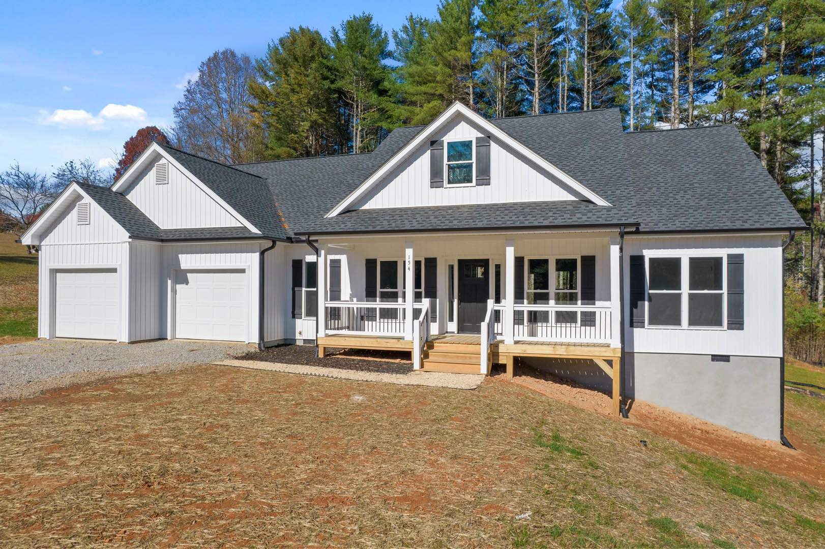 Two-story house with white-framed windows, covered porch featuring a wooden bench, brown dirt driveway, wooden deck and stairs, surrounded by mature trees in the background