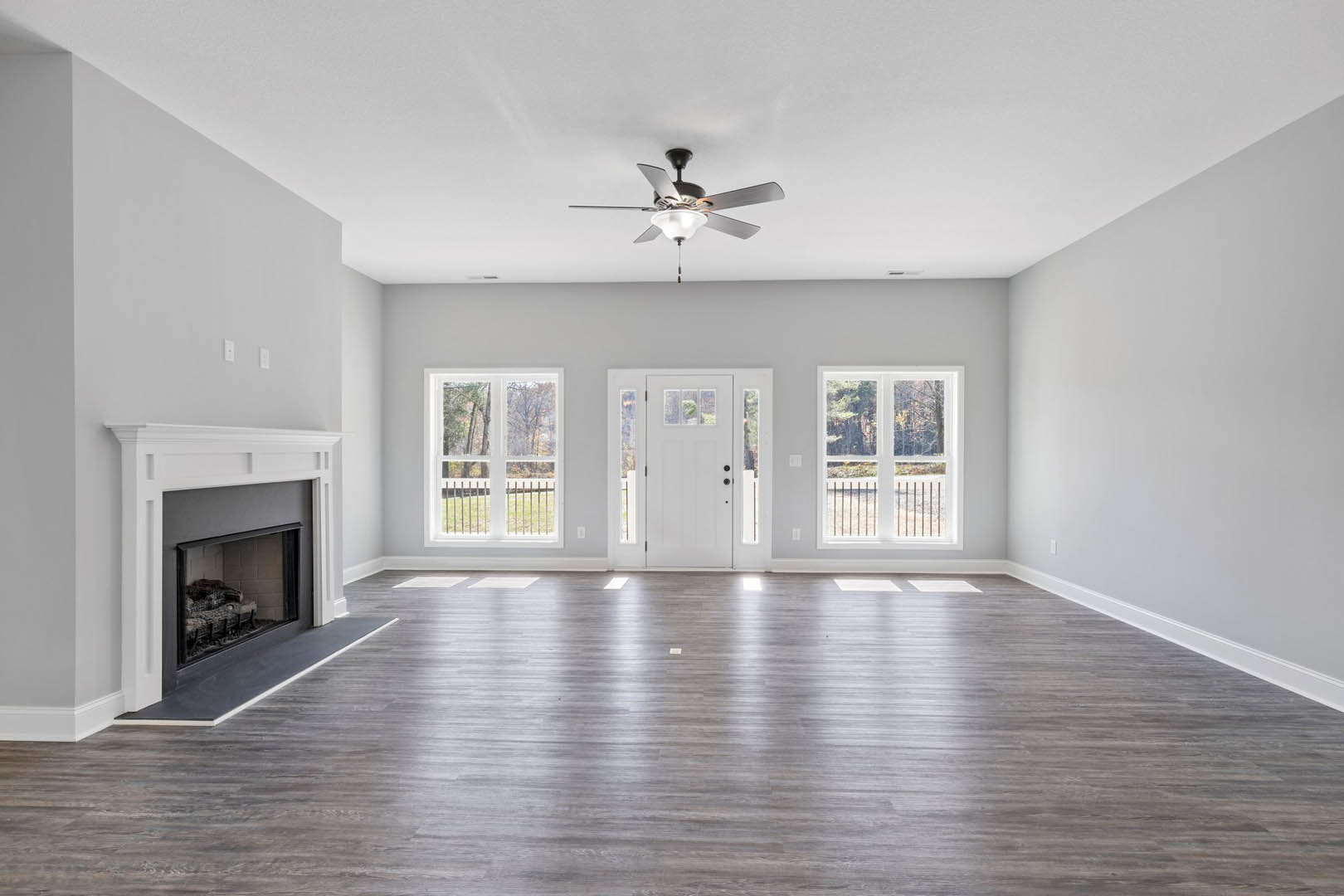 Living room with wood flooring, white walls, ceiling fan with light fixture, white-framed fireplace, window with railing, and white door with glass panes