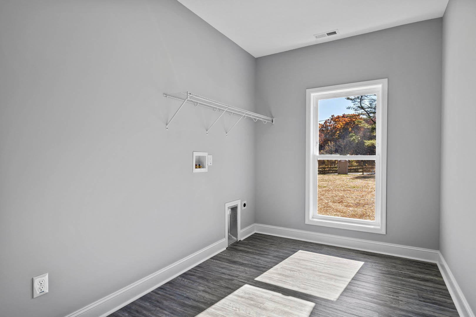 Sunlit room featuring a large window overlooking a yard with trees, wood flooring, white shelf with hooks, wall outlet, and white door with glass panel.