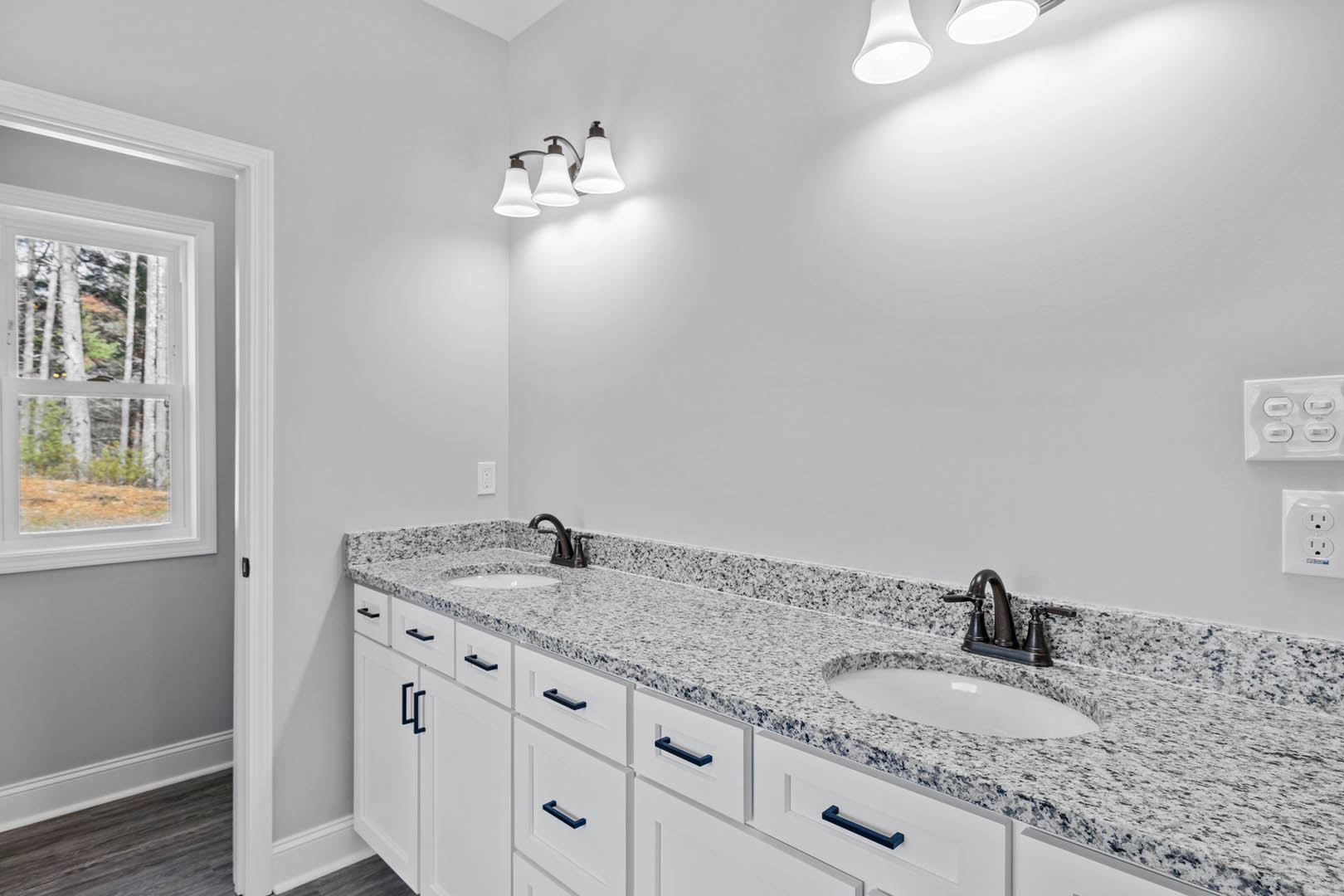 Bathroom featuring white cabinetry, marble countertops, under-mount sink, chrome faucet, three-light fixture, tile backsplash, window with tree view, and white wall outlet