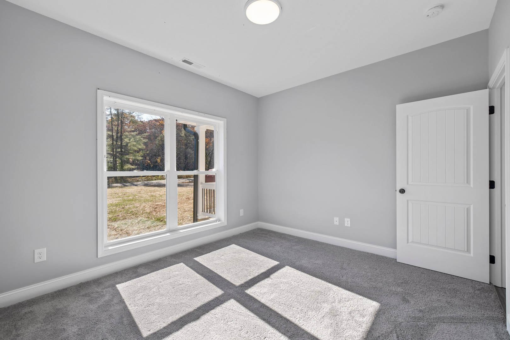 White-walled room featuring a white door with black handle, window overlooking green yard, circular ceiling light, white electrical outlet, and light-colored carpet with subtle