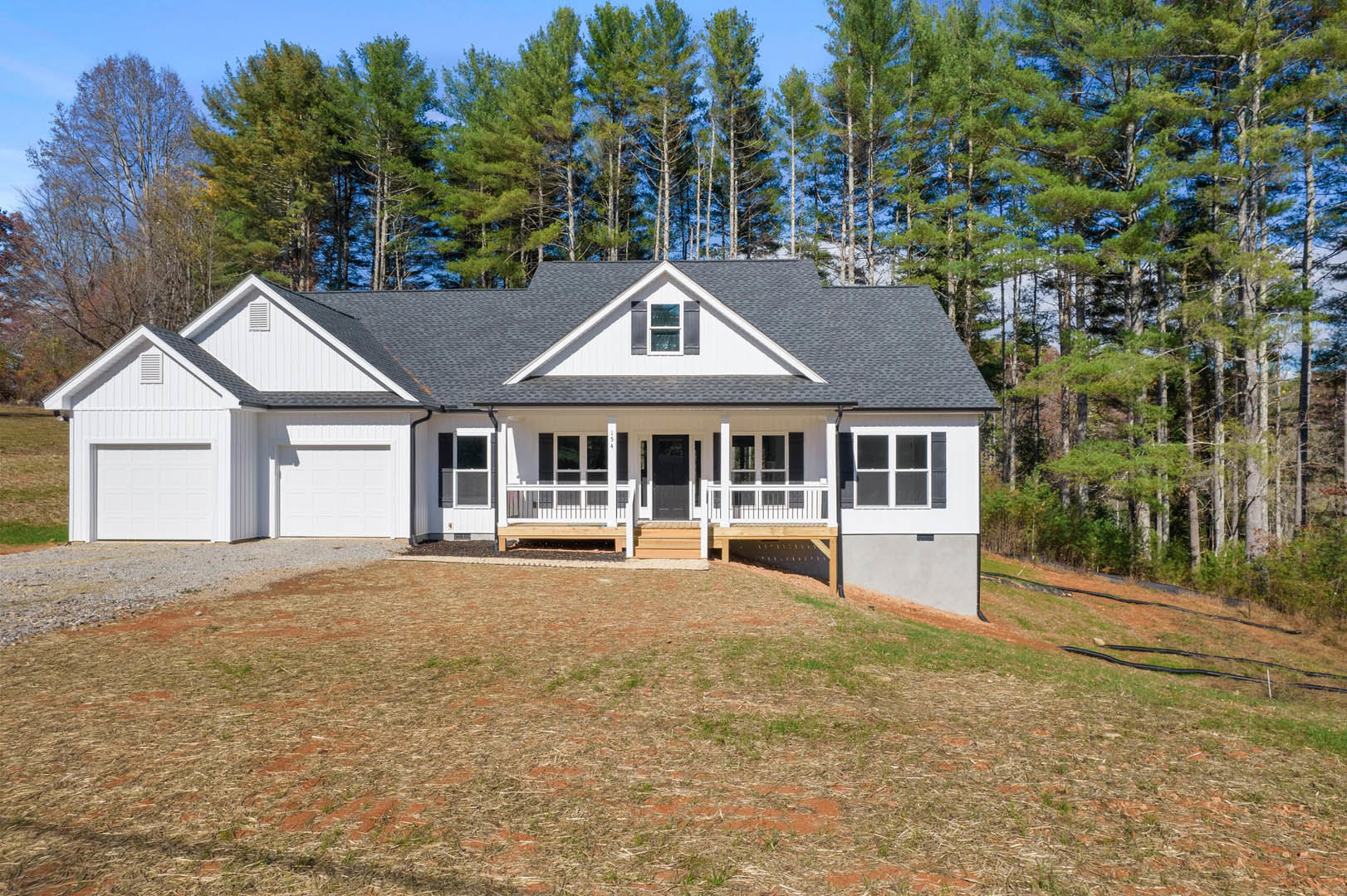 Two-story white house with black door and white trim, attached garage, covered porch, concrete driveway, grassy lawn, mature trees in the background, small cottage visible behind