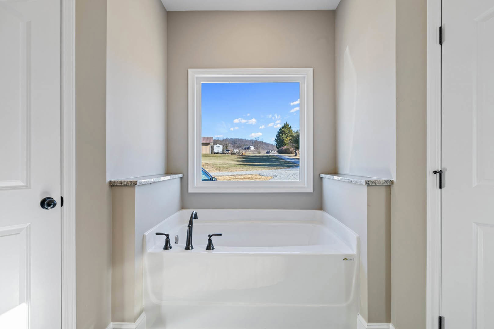 Freestanding white bathtub beneath large window overlooking field and trees, chrome faucet, white walls, black hardware, natural light filling bathroom
