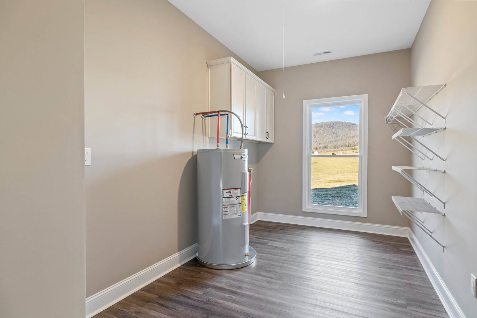 Wood-floored utility room featuring a large grey water heater tank, ceiling vent, exposed pipes, plaster walls, and a window overlooking a grassy field and hill.
