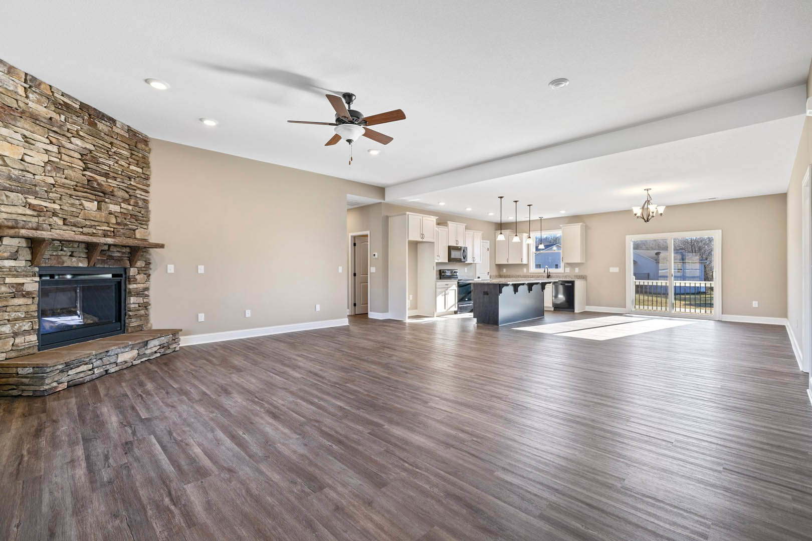 Spacious open floor plan featuring hardwood floors, stone fireplace with glass door, ceiling fan with light fixture, adjacent kitchen area, and large window overlooking fenced yard