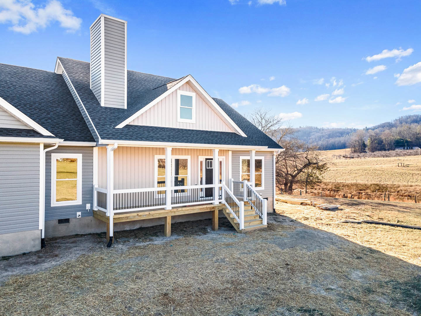 Two-story home with white siding, prominent chimney, covered porch, large windows with white frames, wooden deck with white railing, expansive grassy yard, and cloudy sky overhead