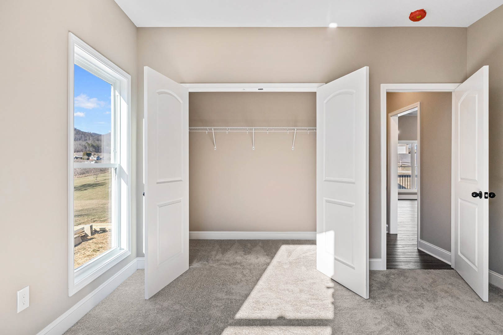 Spacious room with open white doors, closet, white electrical outlet on wall, window overlooking field and neighboring house, light-colored flooring, red object with yellow label
