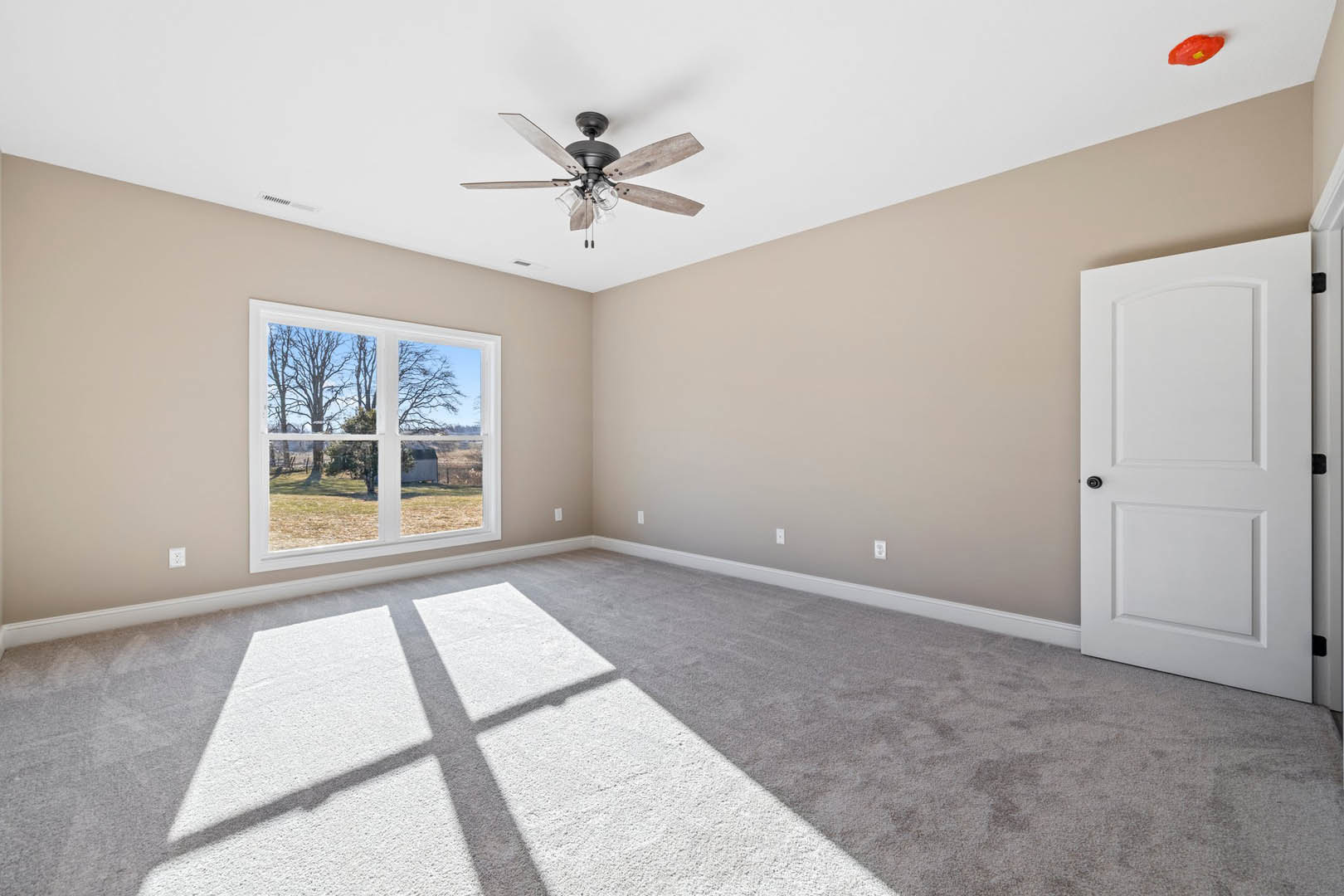 Bedroom with light wood flooring, white plaster walls, ceiling fan with glass light fixtures, large window overlooking farm and trees, white door with black handle, sunlight