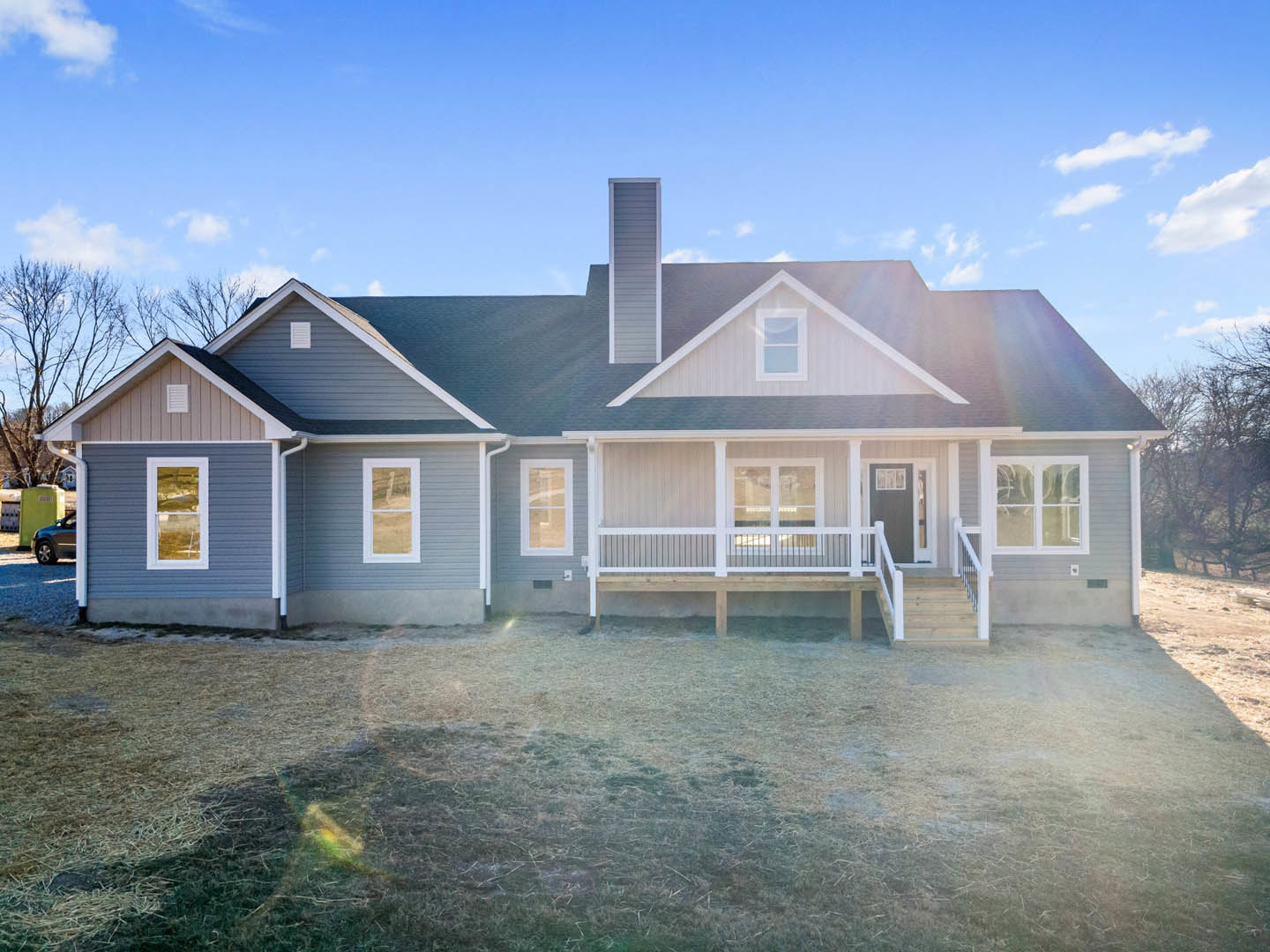 Two-story house with white siding, covered front porch, large windows with white frames, manicured lawn, wooden deck, leafy trees, and clear blue sky
