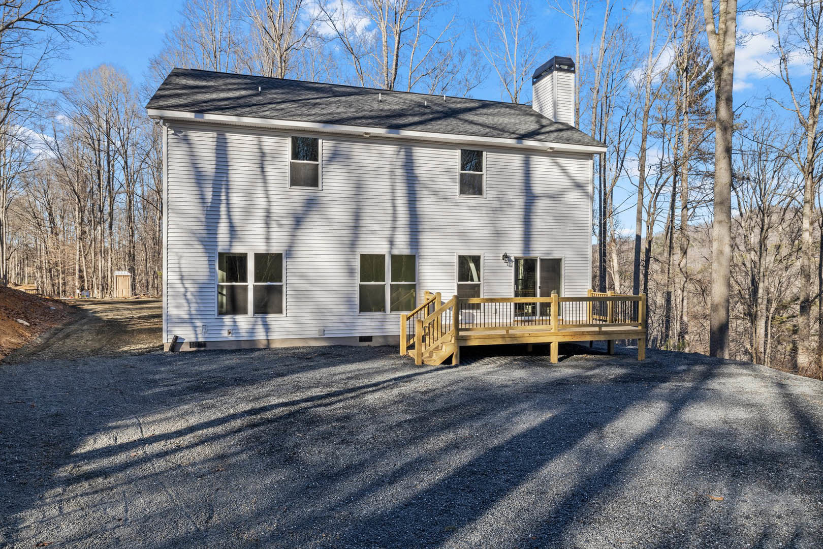 Two-story house with gray siding, white-trimmed windows, elevated wooden deck with stairs, gravel driveway, and wooden bench in front; leafless trees and winter sky in background.