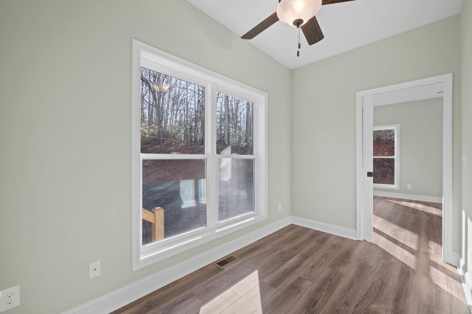 Living room with laminate flooring, white walls, ceiling fan with light, exposed wooden beam, open door, and windows overlooking trees