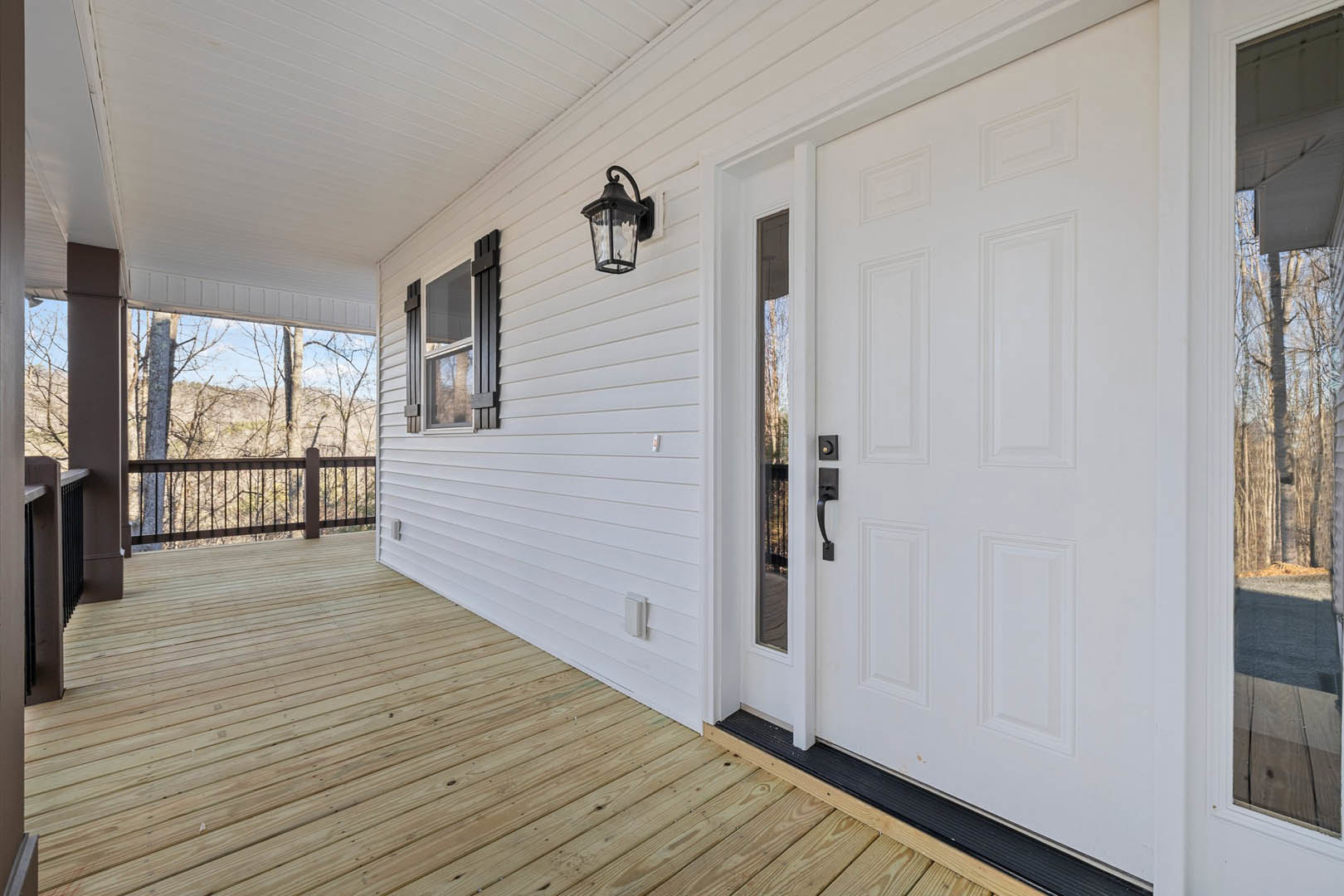 White front door with black handle set in a white wall, wooden deck flooring, metal railing, and exterior porch lamp visible