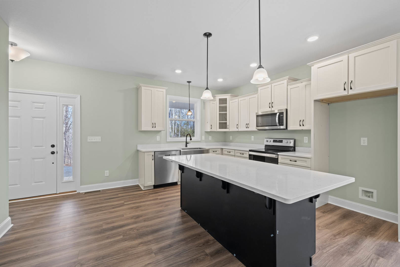 White kitchen cabinets and black island with white countertop, stainless steel sink, built-in microwave, tile backsplash, pendant lighting, person standing near window.