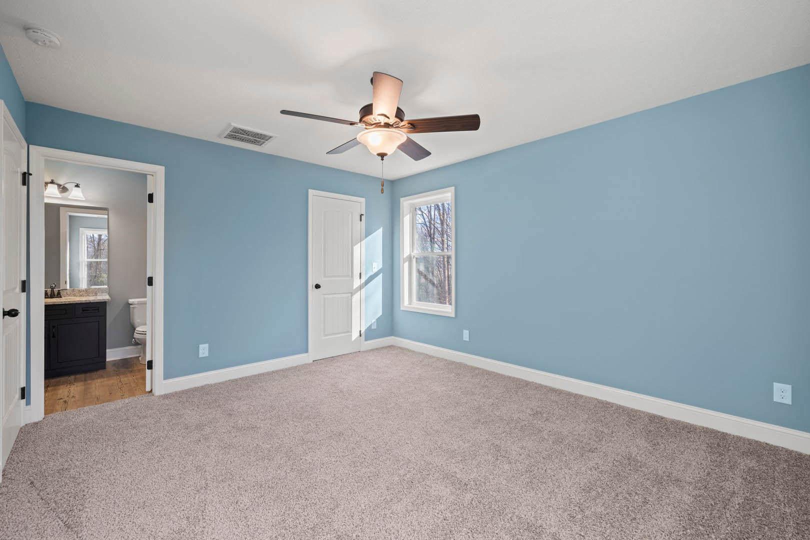 Carpeted room featuring a ceiling fan with light fixture, light blue walls, white door with black knob, window overlooking trees, and black cabinet topped with white countertop