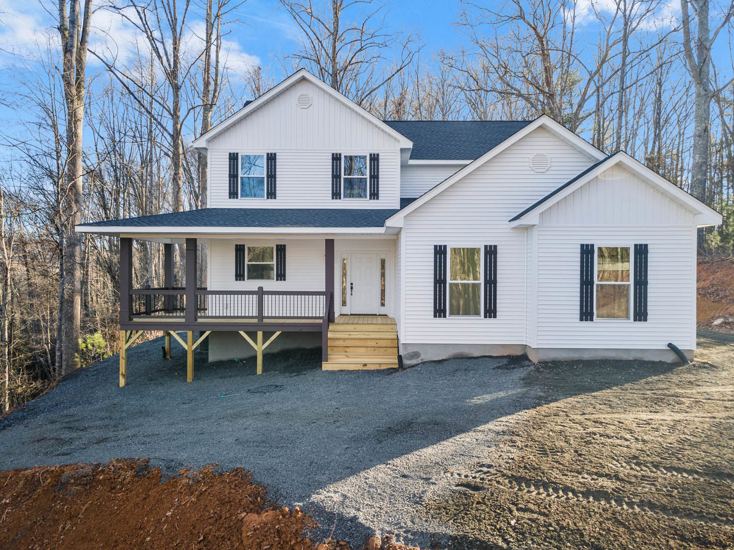 White siding house with covered porch, wooden deck and railing, gravel driveway, white-framed windows, and landscaped ground.