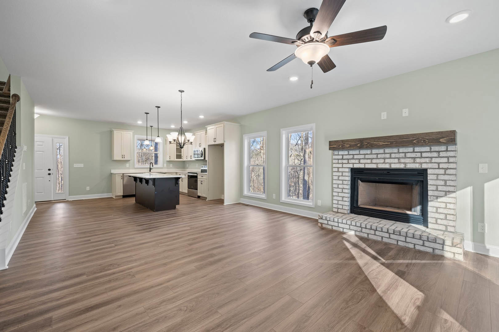 Living room with hardwood floors, glass-door fireplace, ceiling fan with light, black and white rectangular table, open kitchen, large window overlooking trees