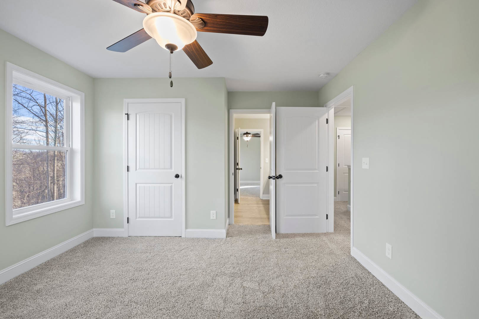 Room with carpeted walls, white doors with black knobs, ceiling fan with light, window showing trees outside, and close-up of light fixture