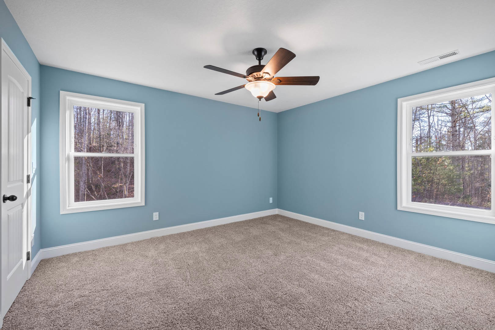 Neutral-toned carpeted room with white walls, ceiling fan with integrated light, large windows showing green trees outside