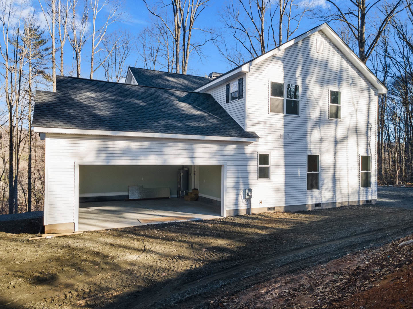 Gray siding house with attached garage, white-framed windows, gable roof, dirt patch in front yard, and trees in background.