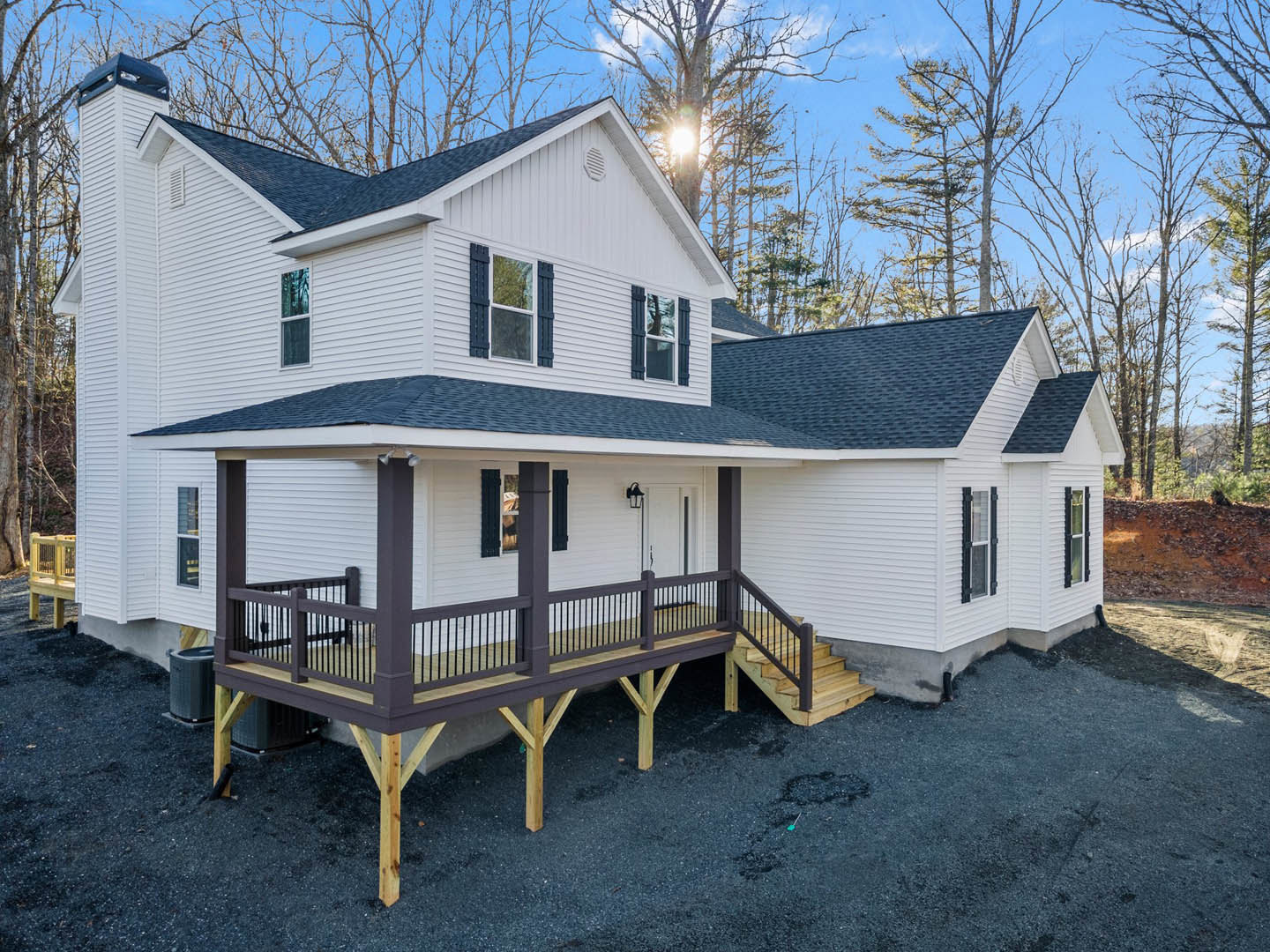 White siding house with covered porch, wooden deck railing, concrete driveway, large windows reflecting nearby trees, and landscaped front yard.